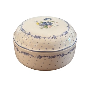 Puede incluir: Una caja redonda de cerámica con tapa. La caja es de color blanco roto con pequeños lunares azules y un diseño floral azul en el borde. La tapa presenta un arreglo floral en azul, rosa y amarillo.