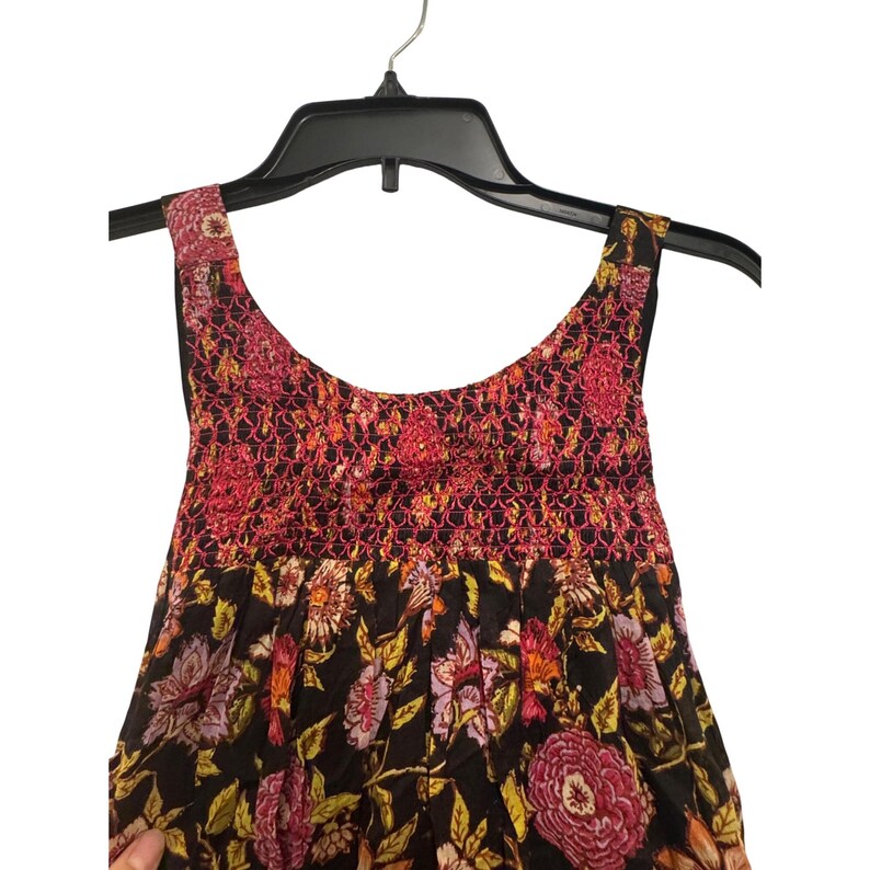 May include: A sleeveless dress with a floral pattern in shades of pink, yellow, and orange on a black background. The bodice features a smocked design in pink and red. The dress is hanging on a black hanger.