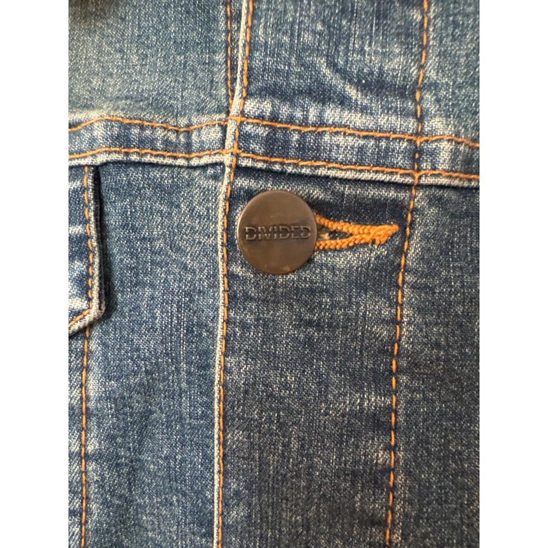 May include: Close-up of a blue denim jacket with a brown button that says "DIVIDED" in white lettering.