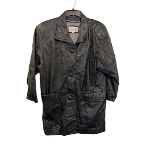 Könnte beinhalten: Eine schwarze Lederjacke mit spitzem Kragen, drei Knöpfen auf der Vorderseite und zwei großen aufgesetzten Taschen. Das Innenetikett trägt die Aufschrift Pelle Studio. Die Jacke hat lange Ärmel und eine strukturierte Passform, hängend an einem schwarzen Kunststoffkleiderbügel.