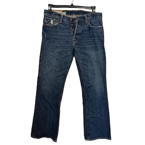 Pode incluir: Jeans de ganga de lavagem escura com fecho de botões. Os jeans têm um design clássico de cinco bolsos e uma ligeira abertura da perna em bota. Os jeans estão pendurados num cabide preto.