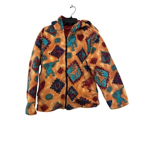 Peut inclure: Veste polaire à capuche orange avec un motif du Sud-Ouest. Le motif répète des figures turquoise et rouges et des formes géométriques. La veste a une fermeture éclair intégrale et une doublure intérieure rouge.