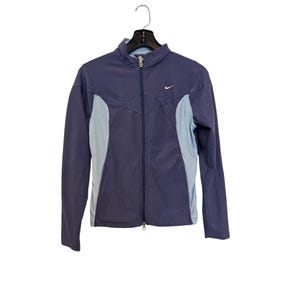 Może przedstawiać: Granatowa kurtka dresowa Nike z jasnoniebieskimi panelami bocznymi i pełnym zapięciem na zamek. Kurtka ma małe logo Nike na piersi. Wisi na czarnym wieszaku na białym tle.