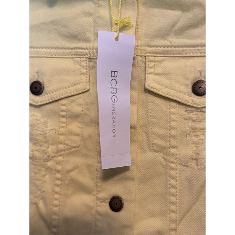 May include: Pale yellow denim jacket with distressed detailing and dark brown buttons. The jacket features two chest pockets and a tag that reads "BCBGGENERATION".