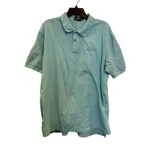 May include: A light blue polo shirt with short sleeves, a collar, and a three-button placket. The shirt features a small, embroidered logo on the chest. The shirt is hanging on a black hanger.
