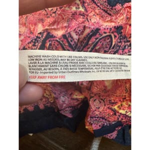 May include: Close-up of a fabric tag with care instructions. The tag is white with black text, including washing instructions and a warning to keep away from fire. The fabric has a paisley pattern in shades of pink, red, and black.