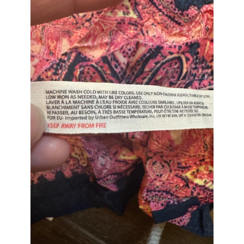 May include: Close-up of a fabric tag with care instructions. The tag is white with black text, including washing instructions and a warning to keep away from fire. The fabric has a paisley pattern in shades of pink, red, and black.