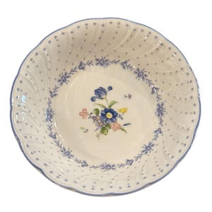 Könnte beinhalten: Weißer Keramikschüssel mit blauem Blumendesign. Die Schüssel hat einen gewellten Rand und ein Muster aus blauen Punkten. Eine zentrale Blumenanordnung umfasst blaue, rosa und gelbe Blumen. Der Rand ist blau umrandet.