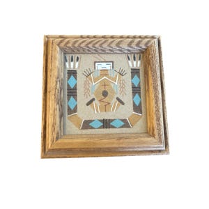 Op de afbeelding: Een ingelijst zand schilderij dat een indiaanse figuur in traditionele kleding met turquoise en bruine accenten afbeeldt. Het schilderij is op een beige achtergrond en ingelijst in een natuurlijke houten lijst.