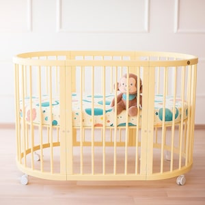 May include: A light yellow oval crib with vertical bars and a patterned mattress featuring whales and other sea creatures. A brown stuffed monkey with a blue bib sits inside the crib. The crib has wheels.