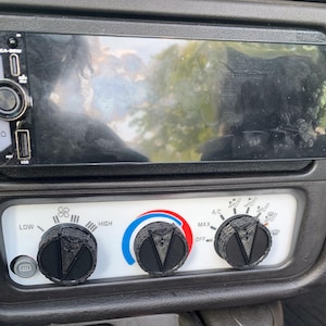 Op de afbeelding: Een zwarte dashboardconsole van een auto met een rechthoekig digitaal mediascherm boven een wit paneel voor klimaatregeling. Het paneel bevat drie zwarte, getextureerde ronde knoppen met driehoekige indicatoren en een kleine grijze knop.