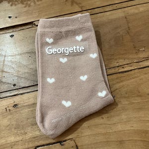 May include: A pair of blush-colored socks with white heart patterns. The word "Georgette" is printed in white across the top of the socks. The socks are folded and resting on a wooden surface.