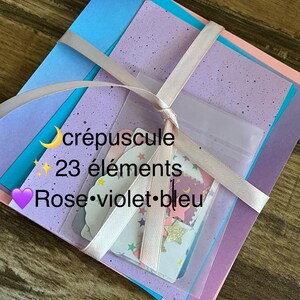 Peut inclure: Un ensemble de feuilles carrées en bleu, lilas moucheté et rose pâle, noué par un ruban rose clair. Une pochette transparente contient 23 formes découpées, dont des étoiles et des nuages. Le texte indique : crépuscule 23 éléments Rose violet bleu.