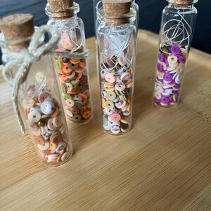 Peut inclure: Plusieurs petits flacons en verre avec des bouchons en liège sont remplis de perles rondes et colorées. Les perles sont dans des tons orange, rose, vert et blanc. Une bouteille est attachée avec de la ficelle. Les bouteilles sont sur un plateau en bois brun clair.