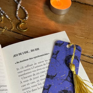 Puede incluir: Un marcapáginas azul con borla dorada, con diseños de luna creciente y mariposas, descansa sobre un libro abierto. También se ven un collar dorado con un colgante de luna creciente y una vela naranja encendida.