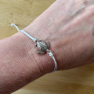May include: A silver turtle charm bracelet with a white cord. The turtle charm is detailed with textured shell and flippers. The bracelet is simple and adjustable, suitable for everyday wear.