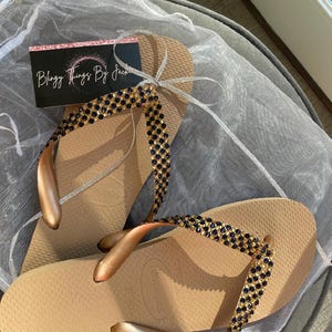 May include: A pair of gold flip flops with a black and gold rhinestone embellishment. The flip flops are on a white background. The text "Blingy Things By Jack" is visible on a pink and white card.