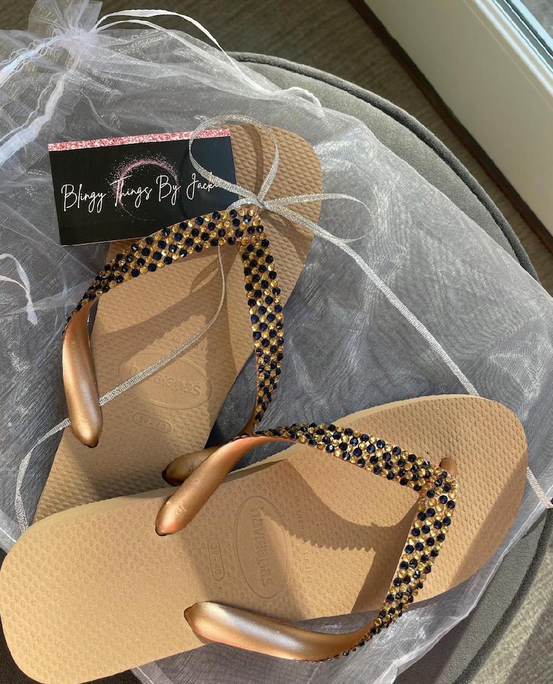 May include: A pair of gold flip flops with a black and gold rhinestone embellishment. The flip flops are on a white background. The text "Blingy Things By Jack" is visible on a pink and white card.