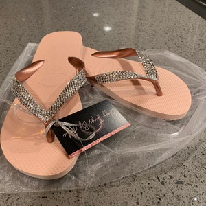 May include: Pale pink Havaianas flip-flops with rose gold straps, adorned with sparkling rhinestones. The flip-flops are presented in a sheer white gift bag, accompanied by a tag reading "Dizzy Things By Sophie".