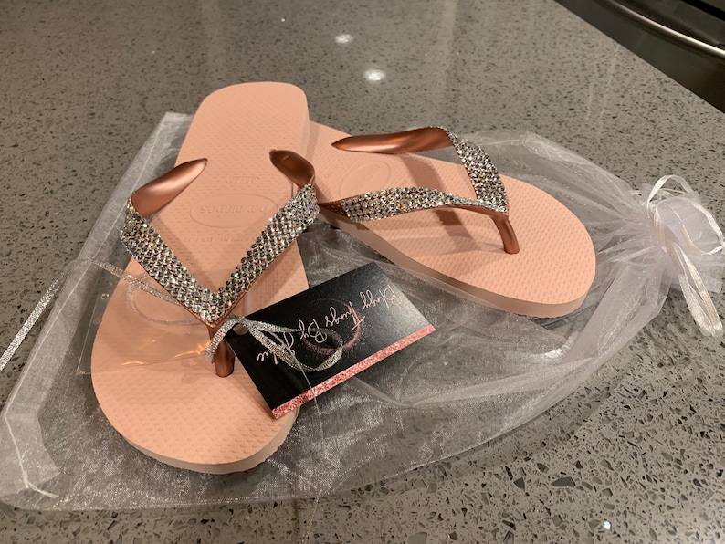May include: Pale pink Havaianas flip-flops with rose gold straps, adorned with sparkling rhinestones. The flip-flops are presented in a sheer white gift bag, accompanied by a tag reading "Dizzy Things By Sophie".