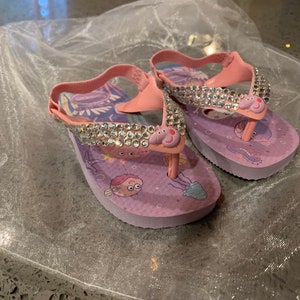 May include: A pair of pink and purple children's flip-flops with a sparkly rhinestone strap. The flip-flops feature a cartoon pig design and sea creature graphics. The straps are pink and the soles are purple.