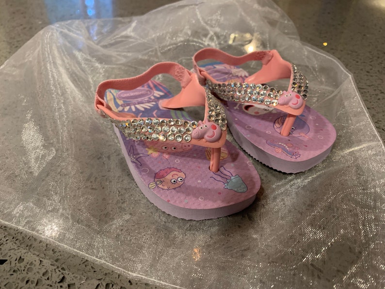 May include: A pair of pink and purple children's flip-flops with a sparkly rhinestone strap. The flip-flops feature a cartoon pig design and sea creature graphics. The straps are pink and the soles are purple.