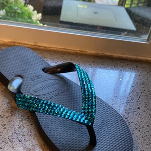 May include: Black flip-flop sandals with a turquoise blue rhinestone strap. The strap has the text "Havaianas" on it.