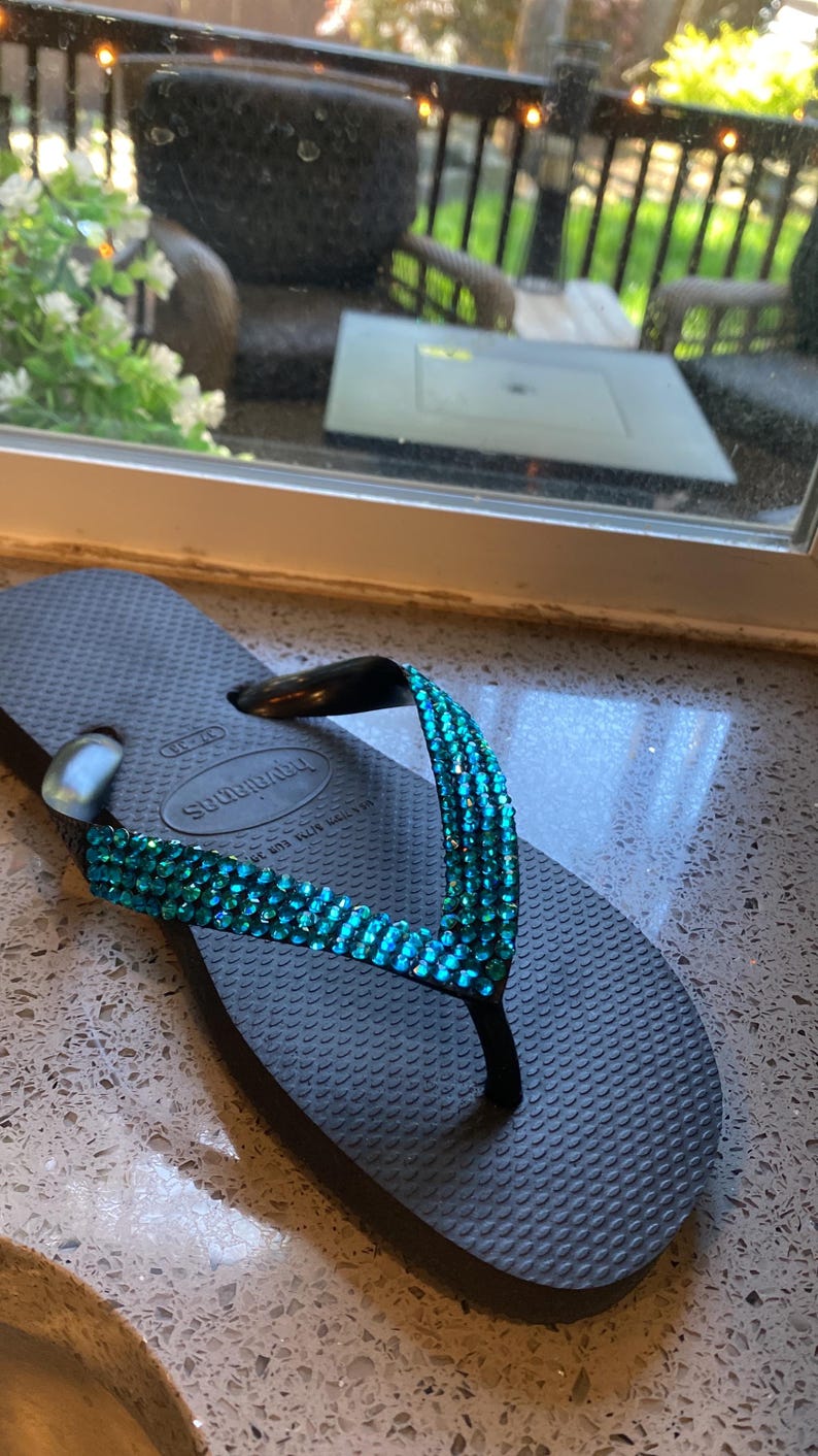 May include: Black flip-flop sandals with a turquoise blue rhinestone strap. The strap has the text "Havaianas" on it.