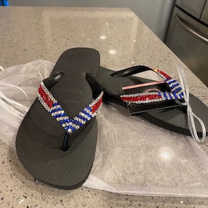 May include: Black flip-flops with red, white, and blue rhinestone straps. The sandals are presented on a white mesh bag. These flip-flops are a summer footwear item.