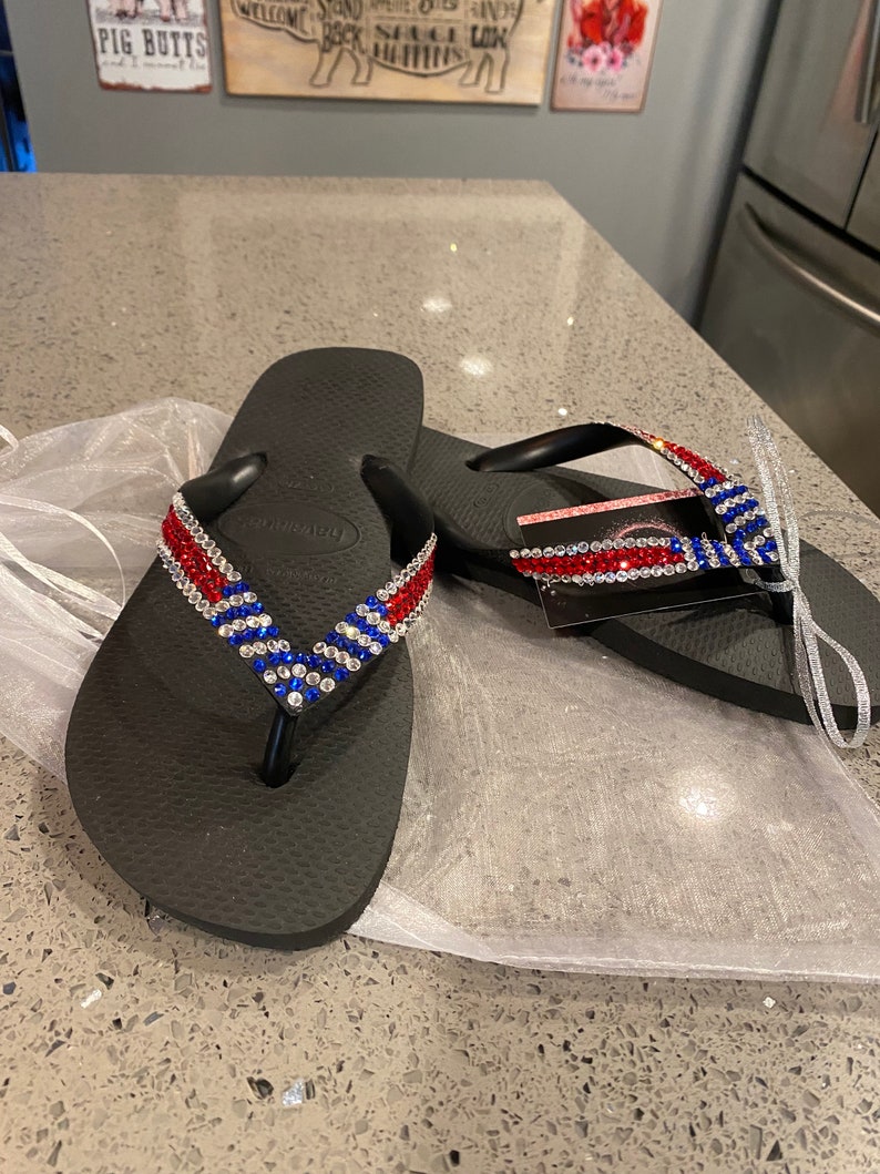 May include: Black flip-flops with red, white, and blue rhinestone straps. The sandals are presented on a white mesh bag. These flip-flops are a summer footwear item.