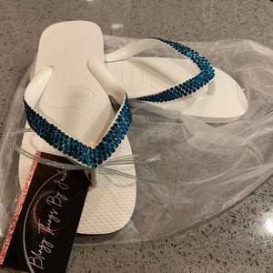 May include: White flip flops with a blue and green rhinestone design. The flip flops are in a white tulle bag with a pink and white business card that says "Blingy Things By Jackie".