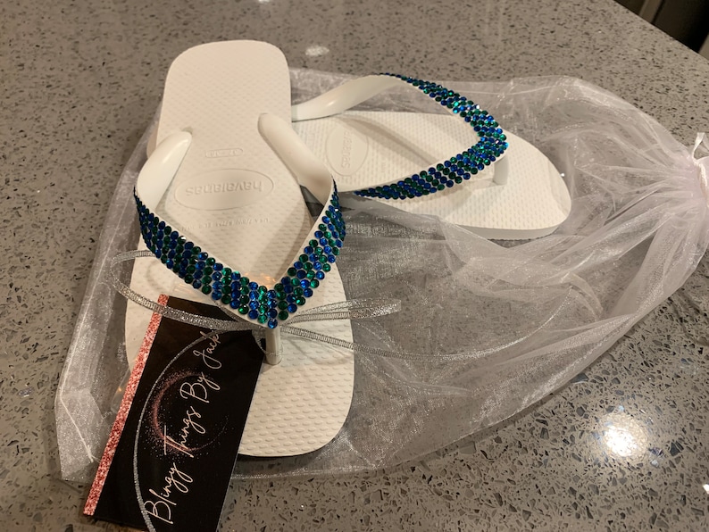 May include: White flip flops with a blue and green rhinestone design. The flip flops are in a white tulle bag with a pink and white business card that says "Blingy Things By Jackie".