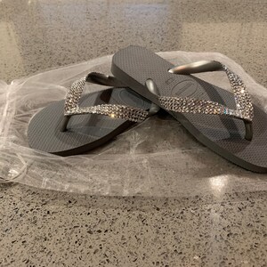 May include: Grey flip-flops with sparkling rhinestone straps. The sandals are resting on a white, translucent material. These flip-flops are a casual footwear choice, ideal for warm weather or beach outings.