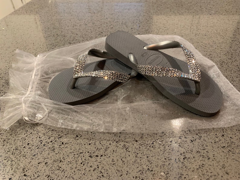 May include: Grey flip-flops with sparkling rhinestone straps. The sandals are resting on a white, translucent material. These flip-flops are a casual footwear choice, ideal for warm weather or beach outings.