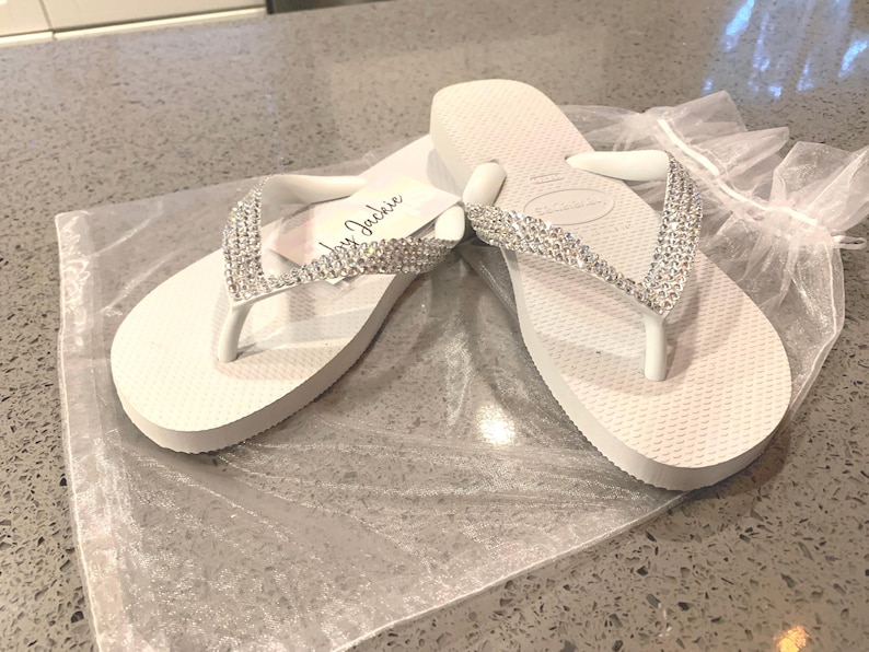 May include: White flip-flops with rhinestone straps. The sandals are on a clear plastic bag. The text "by Jackie" is on a small white tag attached to one of the flip-flops. The flip-flops are on a grey surface.