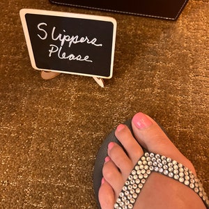 May include: A foot wearing a black flip-flop with a strap adorned with clear rhinestones. The toenails are painted pink. A small chalkboard sign reads "Slippers Please".