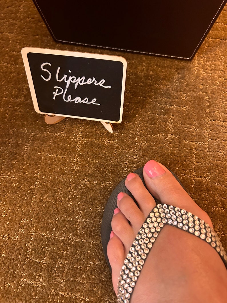 May include: A foot wearing a black flip-flop with a strap adorned with clear rhinestones. The toenails are painted pink. A small chalkboard sign reads "Slippers Please".
