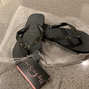 May include: A pair of black Havaianas flip-flops with a sparkling, jeweled strap. The flip-flops are presented on a white, translucent bag, with a tag that reads "Bloggy Things By Jackie". These flip-flops are a fashionable footwear choice.