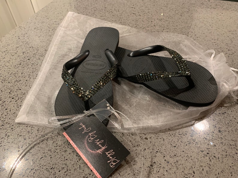 May include: A pair of black Havaianas flip-flops with a sparkling, jeweled strap. The flip-flops are presented on a white, translucent bag, with a tag that reads "Bloggy Things By Jackie". These flip-flops are a fashionable footwear choice.