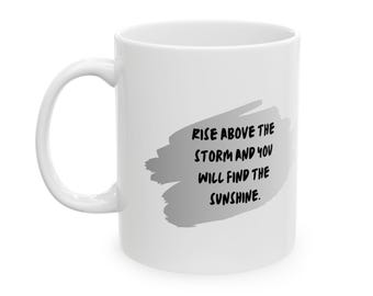 May include: A white ceramic mug featuring a gray brushstroke graphic on the side. The text printed in black over the gray shape reads: RISE ABOVE THE STORM AND YOU WILL FIND THE SUNSHINE. The mug has a curved handle on the left side.