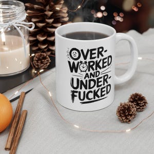 May include: White ceramic mug with a black handle, filled with a dark liquid. The mug features the text "OVER WORKED AND UNDER FUCKED" in a bold, black font. The design includes a clock and a frowning face.