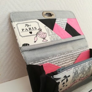 Puede incluir: Una cartera de tela negra y gris con cierre de cremallera. La cartera tiene un forro de tela estampado en rosa y negro y una solapa de tela blanca con el texto "Moi Paris" y un pequeño corazón.
