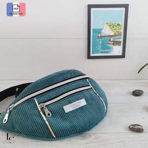 May include: Teal corduroy fanny pack with two zipped compartments and a black strap. The bag has white and silver accents, and a small label that reads "LOUMJANNE Créations". A framed print of a coastal scene hangs in the background.