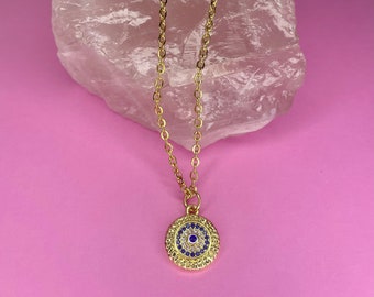 Puede incluir: Collar de cadena dorada con un colgante redondo azul de ojo turco. El colgante está rodeado de pequeñas piedras transparentes y azules.
