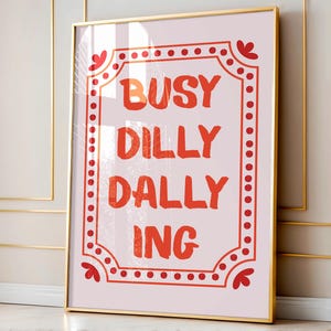 May include: A framed art print with the words "BUSY DILLY DALLY ING" in orange lettering on a light pink background. The text is framed by a red dotted border with decorative corner elements. The frame is gold.