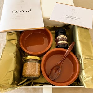 May include: A gift box containing two terracotta ramekins, a 60 ml jar of Nielsen-Massey vanilla bean paste, a small glass jar of brown sugar, a wooden spoon, a recipe booklet titled Custard, and a card reading For the person who takes custard seriously.