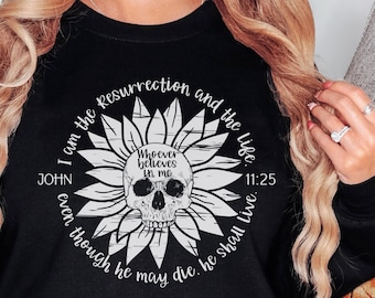 May include: Black sweatshirt featuring a white sunflower design with a skull in the center. The design includes the text "I am the resurrection and the life" and "Whoever believes in me" with a Bible verse reference. The sweatshirt is a crew neck.
