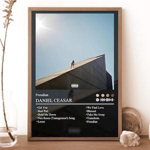 May include: A framed rectangular poster featuring the album cover for Freudian by Daniel Caesar. The artwork shows a person climbing a large, angular concrete structure under a bright sky. Below the image, the tracklist is printed in white text.
