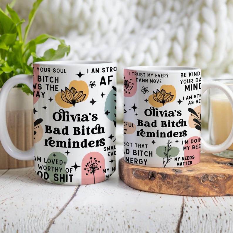 May include: Two white ceramic mugs with the text "Olivia's Bad Bitch Reminders" in black script. The mugs feature floral and star designs in pastel colors. One mug has the text "Your Soul Bitch Wins the Day".