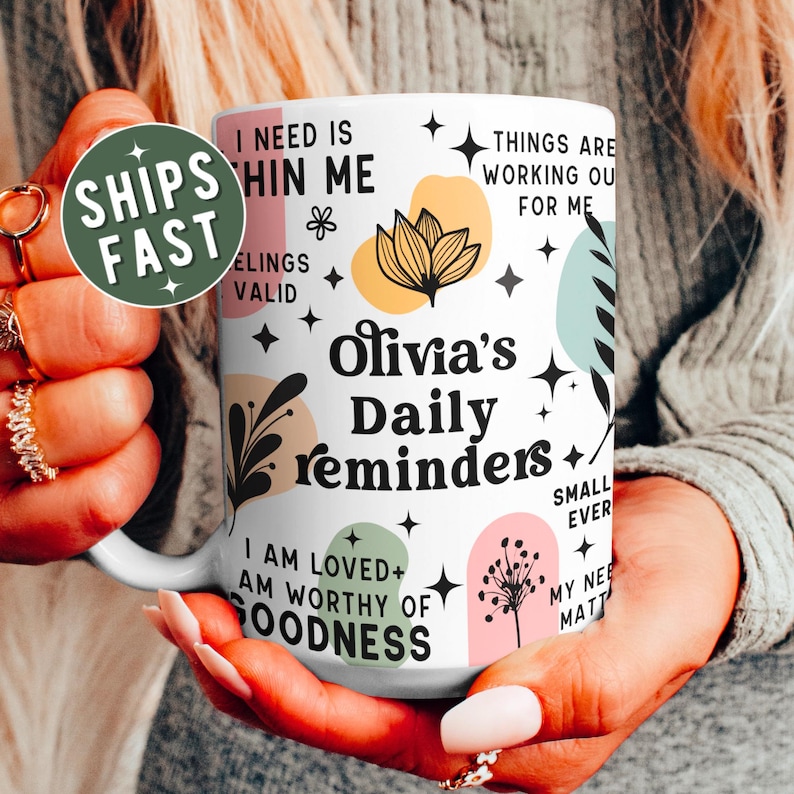 May include: White ceramic mug with the text "Olivia's Daily reminders" in black script. The mug features colorful floral and geometric designs, with phrases like "I am loved" and "Things are working out for me".